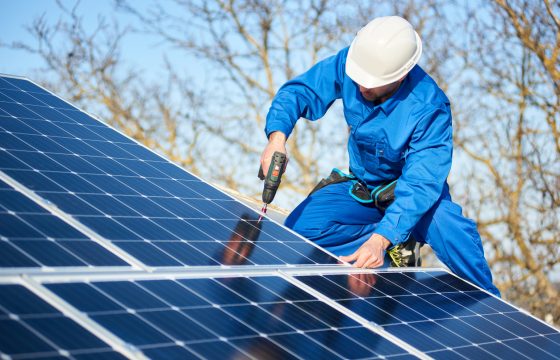 Male engineer in blue suit and protective helmet installing photovoltaic panel system using screwdriver. Professional electrician mounting solar module on roof. Alternative energy ecological concept.